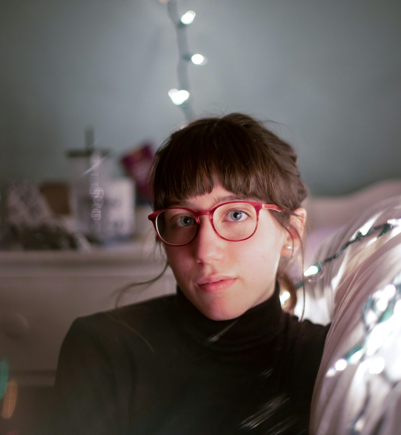 Young person with brown hair and red glasses looks plaintively at the camera. They are sitting down next to their bed, with the bokeh of colourful lights around them.