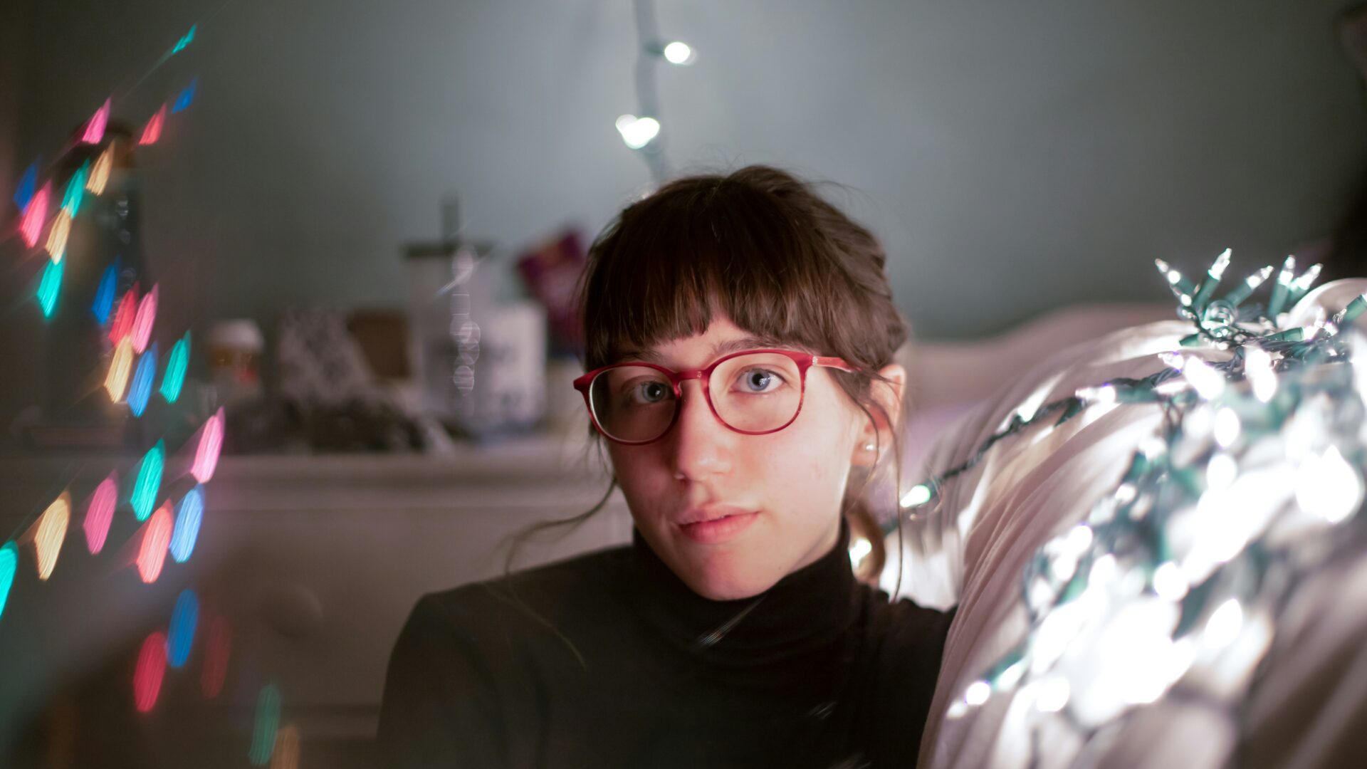 Young person with brown hair and red glasses looks plaintively at the camera. They are sitting down next to their bed, with the bokeh of colourful lights around them.